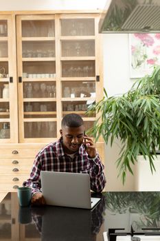 Front view of African-american man talking on mobile phone while using laptop on worktop in kitchen at comfortable home. Authentic home lifestyle setting with young African American male
