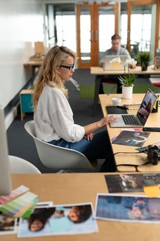 Side view of Caucasian female graphic designer using laptop at desk in office. Caucasian executive man sitting with laptop in the background. This is a casual creative start-up business office for a diverse team