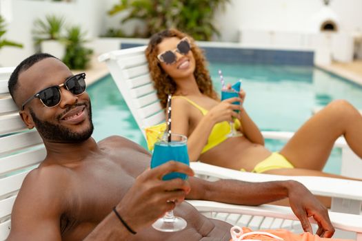 Front view of happy diverse couple having cocktail drink while relaxing on a sun lounger near swimming pool. Summer fun at home by the swimming pool