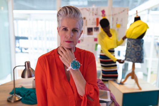 Portrait of mature Caucasian female fashion designer standing in design studio. African american coworker working on dress in the background. This is a casual creative start-up business office for a diverse team
