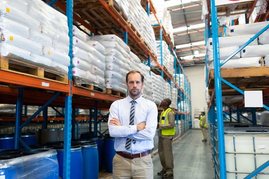 Front view of handsome mature Caucasian male supervisor standing with arms crossed and looking at camera in warehouse. African-american colleagues working behind him. This is a freight transportation and distribution warehouse. Industrial and industrial workers concept