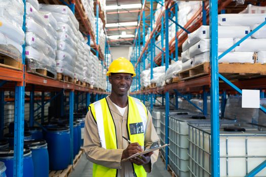 Portrait happy mature African-american male worker looking at camera while writing on clipboard in warehouse. This is a freight transportation and distribution warehouse. Industrial and industrial workers concept