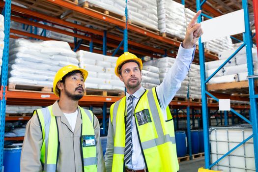 Front view of handsome mature Caucasian male supervisor standing with Asian worker and pointing at distance in warehouse. This is a freight transportation and distribution warehouse. Industrial and industrial workers concept