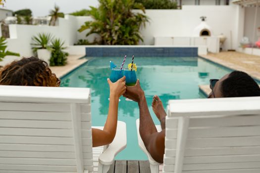 Rear view of cute diverse couple toasting glasses of cocktail while relaxing on a sun lounger near swimming pool in the backyard. Summer fun at home by the swimming pool