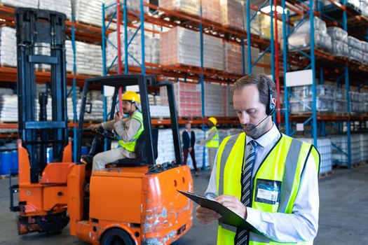 Front view of handsome Caucasian male supervisor with headset writing on clipboard in warehouse. This is a freight transportation and distribution warehouse. Industrial and industrial workers concept