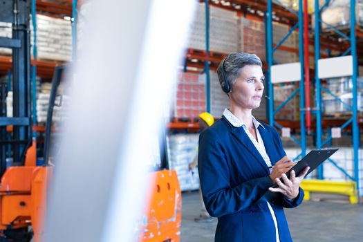 Side view of thoughtful mature Caucasian female manager with headset and clipboard looking away in warehouse. This is a freight transportation and distribution warehouse. Industrial and industrial workers concept