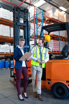 Front view of mature Caucasian male and female staff working together near forklift in warehouse. This is a freight transportation and distribution warehouse. Industrial and industrial workers concept