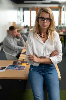 Portrait of Caucasian female graphic designer sitting with arms crossed at desk in office. Caucasian executive man working in the background. This is a casual creative start-up business office for a diverse team