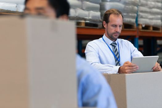 Front view of handsome mature Caucasian male supervisor working on digital tablet in warehouse. On the foreground Asian man holding cardboard boxes. This is a freight transportation and distribution warehouse. Industrial and industrial workers concept