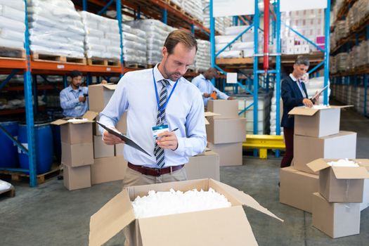 Front view of handsome Caucasian male supervisor checking stocks in warehouse. Diverse warehouse workers unpacking cardboard boxes. This is a freight transportation and distribution warehouse. Industrial and industrial workers concept