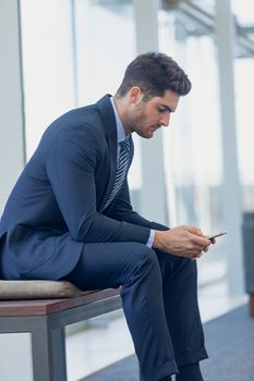 Side view of young Caucasian businessman looking at mobile phone while sitting on bench in modern office. 