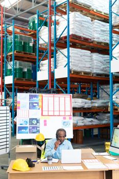 Front view of mature African-american male supervisor talking on headset while using laptop at desk in warehouse. This is a freight transportation and distribution warehouse. Industrial and industrial workers concept