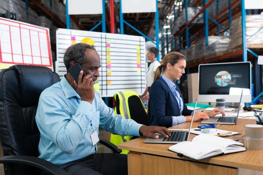 Side view of mature African-american male supervisor using laptop while talking on mobile phone at desk in warehouse. This is a freight transportation and distribution warehouse. Industrial and industrial workers concept