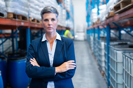 Portrait close-up of beautiful mature Caucasian female manager standing with arm crossed and looking at camera in warehouse. African-american colleague standing in the background. This is a freight transportation and distribution warehouse. Industrial and industrial workers concept