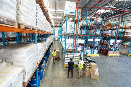 High angle view of diverse warehouse staff working together in warehouse. African-american man is pulling pallet jack with cardboard boxes on it. This is a freight transportation and distribution warehouse. Industrial and industrial workers concept