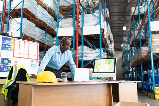 Front view of mature African-american male supervisor working on laptop at desk in warehouse. This is a freight transportation and distribution warehouse. Industrial and industrial workers concept