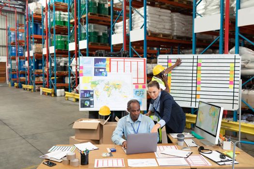 High angle view of mature Caucasian female manager and African-american male supervisor discussing over laptop at desk in warehouse. This is a freight transportation and distribution warehouse. Industrial and industrial workers concept