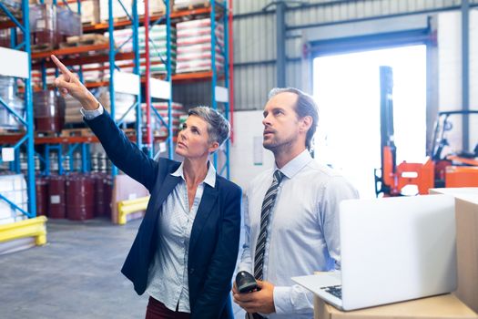 Front view of beautiful mature Caucasian female manager standing with Caucasian male supervisor and pointing at distance in warehouse. This is a freight transportation and distribution warehouse. Industrial and industrial workers concept