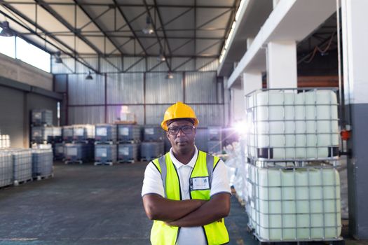 Portrait of African-american male staff in hardhat and reflective jacket standing with arms crossed in warehouse. This is a freight transportation and distribution warehouse. Industrial and industrial workers concept