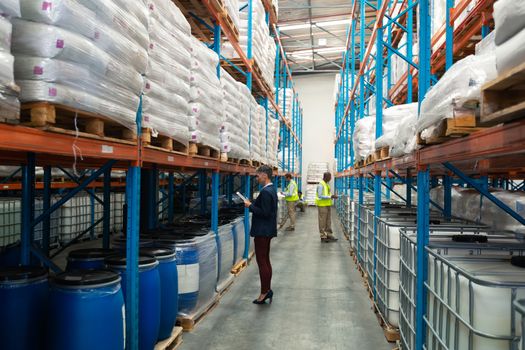 Side view of diverse warehouse staff checking stocks in aisle in warehouse. They are holding clipboards and writing in it. This is a freight transportation and distribution warehouse. Industrial and industrial workers concept