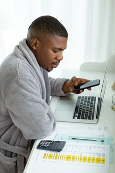 Side view close-up of African-american man looking at bills while using mobile phone on desk in bedroom at comfortable home. Authentic home lifestyle setting with young African American male