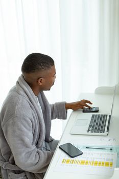 Side view of African-american man calculating bills with calculator on desk in bedroom at comfortable home. Authentic home lifestyle setting with young African American male
