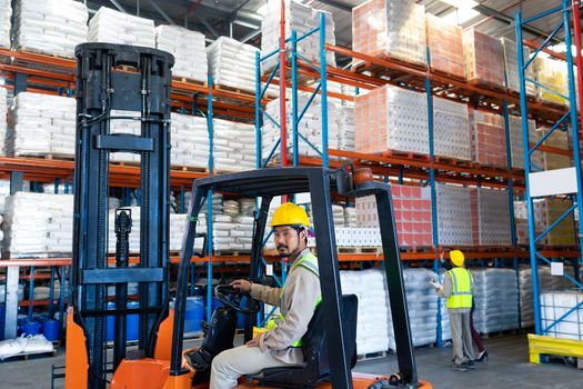 Side view of handsome mature Asian male worker sitting in forklift and looking at camera in warehouse. Diverse colleagues working in the background. This is a freight transportation and distribution warehouse. Industrial and industrial workers concept