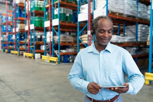 Front view of mature African-american male supervisor with headset using digital tablet in warehouse. This is a freight transportation and distribution warehouse. Industrial and industrial workers concept