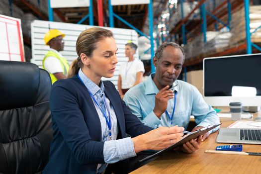 Side view of Caucasian female manager and African-american male supervisor discussing over clipboard at desk in warehouse. This is a freight transportation and distribution warehouse. Industrial and industrial workers concept