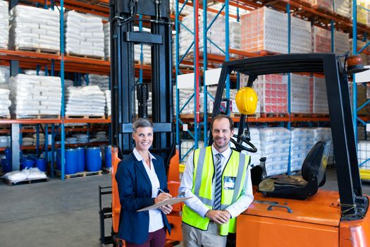 Portrait of happy mature Caucasian male and female staff looking at camera while working together in warehouse. Pretty mature Caucasian woman writing on clipboard in warehouse. This is a freight transportation and distribution warehouse. Industrial and industrial workers concept