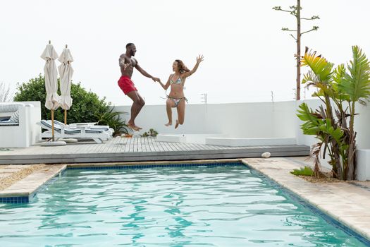 Front view of diverse couple jumping together in swimming pool at the backyard of home. Summer fun at home by the swimming pool