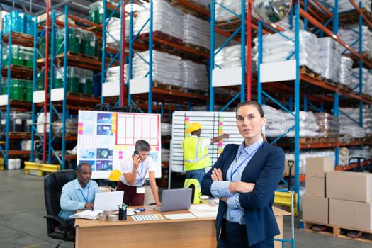 Portrait of confident mature Caucasian female manager standing with arms crossed in warehouse. Diverse colleagues working in the background. This is a freight transportation and distribution warehouse. Industrial and industrial workers concept