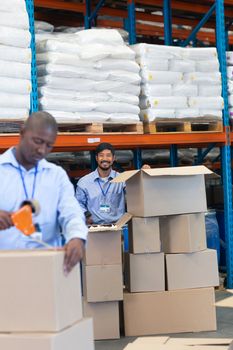 Front view of happy mature Asian male staff looking at camera while checking stock in warehouse. African-american male staff works in front of him. This is a freight transportation and distribution warehouse. Industrial and industrial workers concept