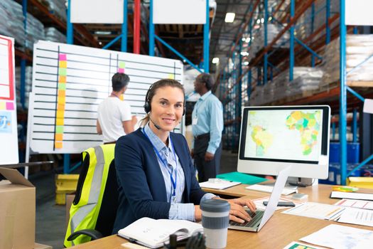 Front view of mature Caucasian female manager with headset using laptop at desk in warehouse. In the background diverse coworkers are discussing in front of whiteboard. This is a freight transportation and distribution warehouse. Industrial and industrial workers concept