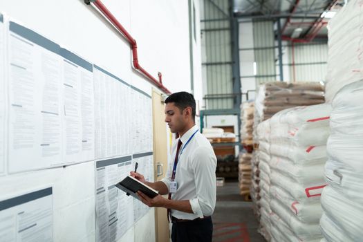 Side view of young Caucasian male supervisor writing on a diary in warehouse. This is a freight transportation and distribution warehouse. Industrial and industrial workers concept