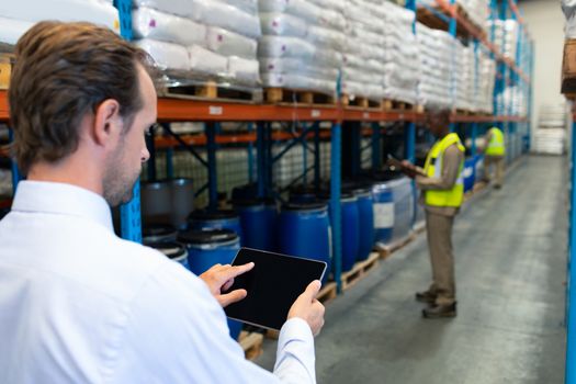 Rear view of mature Caucasian male supervisor working on digital tablet in warehouse. African-american colleagues working in front of him. This is a freight transportation and distribution warehouse. Industrial and industrial workers concept