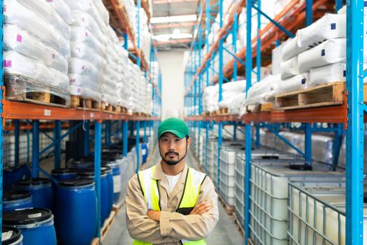 Front view of confident Asian male worker standing with arms crossed and looking at camera in warehouse. This is a freight transportation and distribution warehouse. Industrial and industrial workers concept