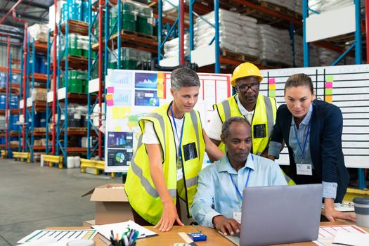 Front view of mature African-american male supervisor with his diverse coworkers discussing over laptop at desk in warehouse. This is a freight transportation and distribution warehouse. Industrial and industrial workers concept