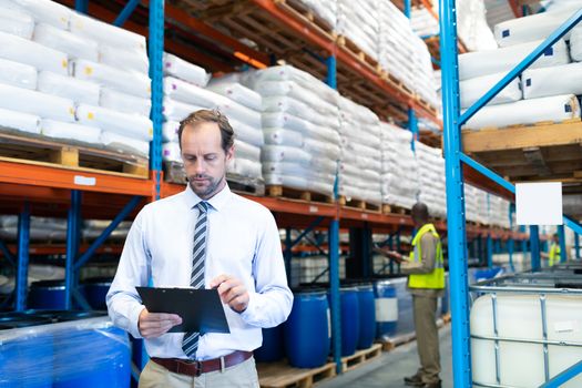 Front view of handsome mature Caucasian male supervisor checking stocks on clipboard in warehouse. African-american colleague is working in the background. This is a freight transportation and distribution warehouse. Industrial and industrial workers concept