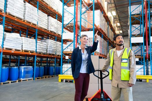 Front view of beautiful mature Caucasian female manager standing with Asian worker and pointing at distance in warehouse. This is a freight transportation and distribution warehouse. Industrial and industrial workers concept