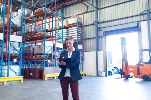 Front view of beautiful mature Caucasian female manager writing on clipboard in warehouse. This is a freight transportation and distribution warehouse. Industrial and industrial workers concept