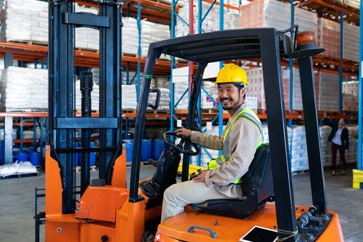 Front view of happy mature Asian male worker sitting in forklift and looking at camera in warehouse. This is a freight transportation and distribution warehouse. Industrial and industrial workers concept