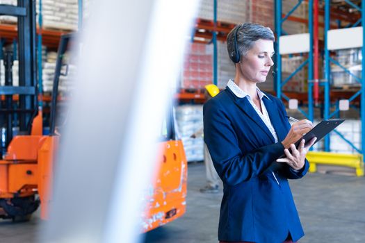 Side view of beautiful mature Caucasian female manager with headset writing on clipboard in warehouse. This is a freight transportation and distribution warehouse. Industrial and industrial workers concept