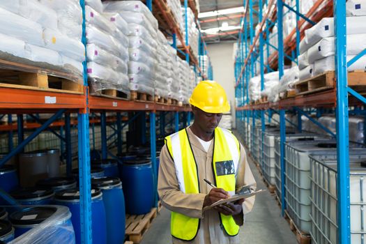 Front view of African-american male worker writing on clipboard in warehouse. This is a freight transportation and distribution warehouse. Industrial and industrial workers concept