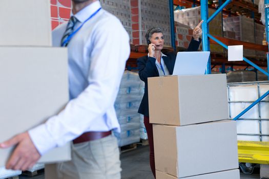 Front view of mature Caucasian female manager with laptop talking on headset in warehouse. On the foreground Caucasian male is holding cardboard boxes. This is a freight transportation and distribution warehouse. Industrial and industrial workers concept