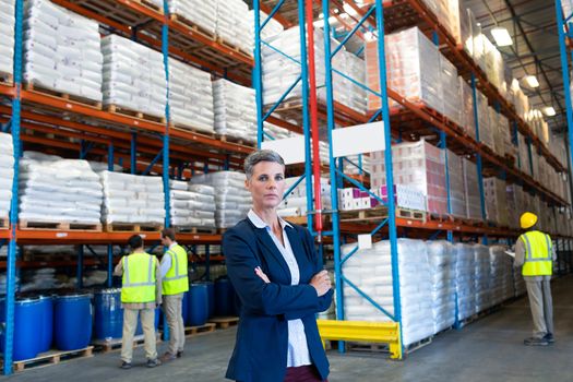 Front view of confident mature Caucasian female manager standing with arms crossed and looking at camera in warehouse. Diverse coworkers standing behind her in aisle. This is a freight transportation and distribution warehouse. Industrial and industrial workers concept