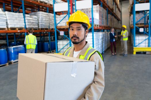 Front view of handsome Asian male worker carrying cardboard box and looking at camera in warehouse. This is a freight transportation and distribution warehouse. Industrial and industrial workers concept