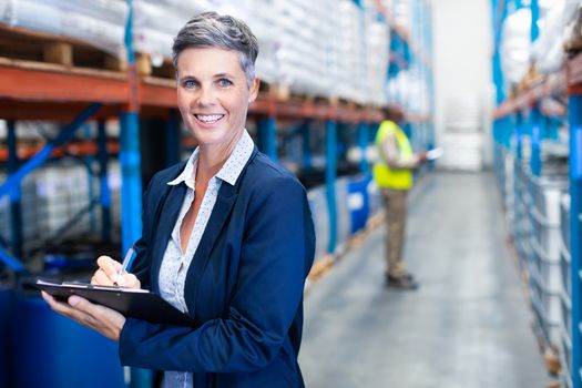 Portrait close-up of beautiful mature Caucasian female manager looking at camera while writing on clipboard in warehouse. African-american colleague standing in the background. This is a freight transportation and distribution warehouse. Industrial and industrial workers concept