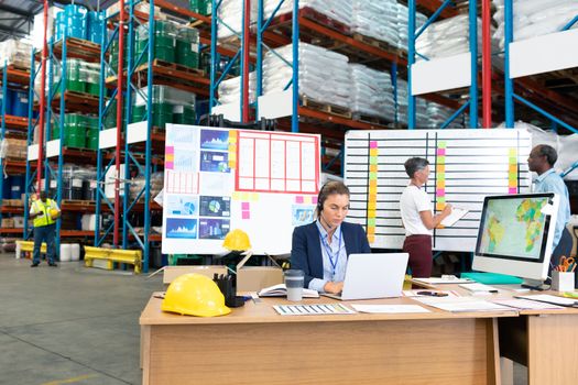 Front view of mature Caucasian female manager with headset using laptop at desk in warehouse. In the background diverse coworkers are discussing in front of whiteboard. This is a freight transportation and distribution warehouse. Industrial and industrial workers concept