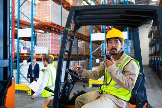 Side view of handsome mature Asian male worker talking on walkie-talkie while driving forklift in warehouse. Diverse colleagues walking in the background. This is a freight transportation and distribution warehouse. Industrial and industrial workers concept